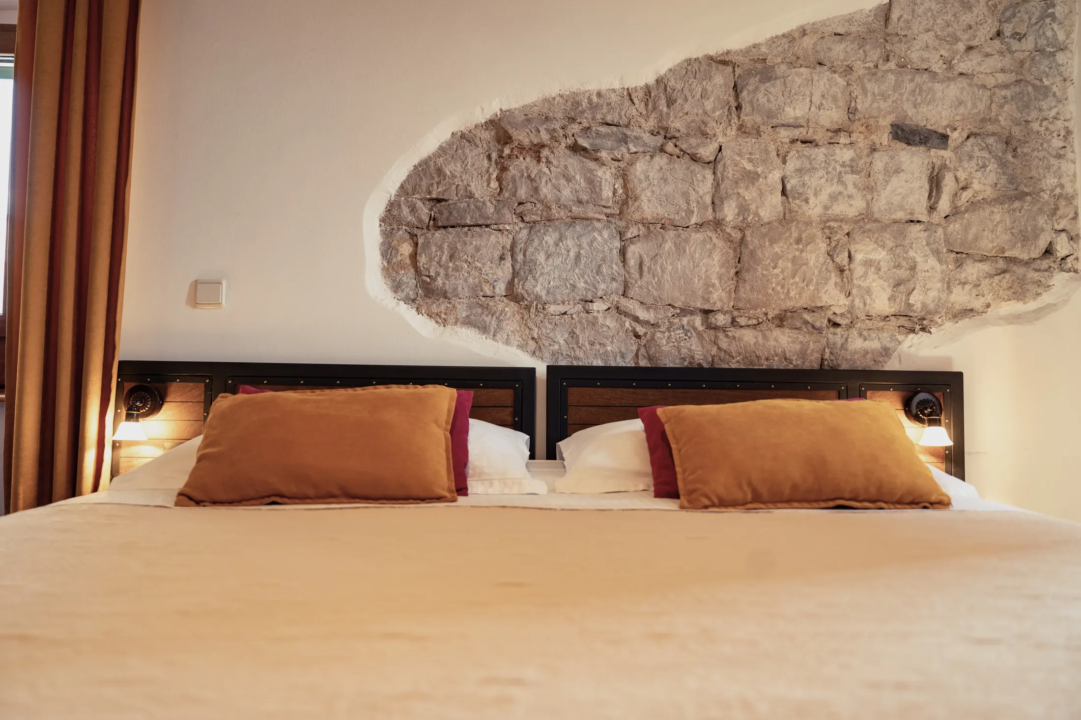 The bed beneath the exposed stone wall, with ochre and burgundy cushions, Hotel Villa Diana, Lučac, Split